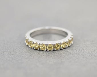 May include: A silver-toned band ring featuring a continuous row of round-cut, pale yellow gemstones set in a prong setting. The metal band has a polished finish, and the stones are arranged evenly around the front half of the circular ring structure.
