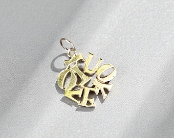May include: A gold-colored pendant with the word "True Love" in an ornate, stylized font. The pendant has a small loop at the top for a chain and is set against a light gray background.