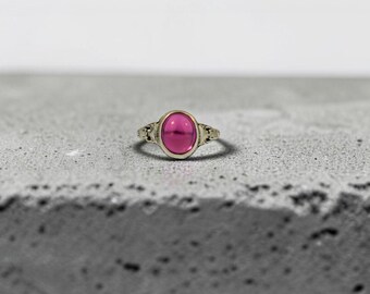 May include: A ring featuring a smooth, oval-shaped, vibrant magenta cabochon stone set in a metallic band. The setting displays intricate, textured detailing on the sides of the band, framing the central gemstone with a polished, reflective finish.