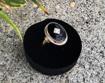 May include: A gold ring featuring an oval black onyx stone set in a textured gold frame. A small, square-cut clear gemstone is embedded in the center of the black onyx. The ring is displayed on a circular black velvet stand.
