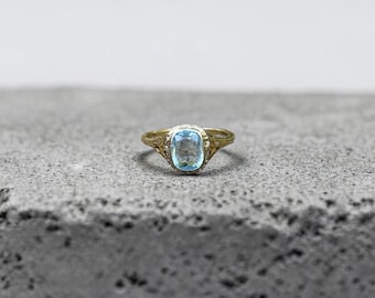 May include: A gold ring featuring a central cushion-cut light blue gemstone. The metal band displays intricate filigree detailing along the sides, leading up to the prong setting that secures the translucent blue stone in a centered position.