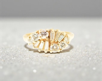 May include: A gold ring featuring a cluster of round brilliant-cut diamonds and tapered baguette-cut diamonds arranged in a fan-like pattern across the top of the band, showcasing a polished yellow gold setting with multiple prong-set stones.