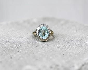 May include: A ring featuring a central oval-cut light blue gemstone set in a decorative metal band. The setting has a textured, filigree-style border surrounding the stone, with a metallic finish that appears to be a combination of silver and gold tones.