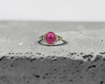 May include: A ring featuring a smooth, oval-shaped, vibrant magenta gemstone set in a metallic band. The metal band displays intricate, textured detailing on the sides, framing the central stone with a polished, reflective finish.