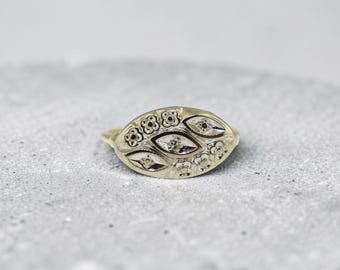 May include: A gold-toned ring featuring an elongated oval face with an engraved pattern of small four-petaled flowers and three central marquise-shaped motifs, each containing a tiny embedded stone at its center point.