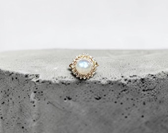 May include: A gold ring featuring a central round iridescent moonstone cabochon surrounded by a halo of small, clear, sparkling stones. The setting is intricate, with a scalloped edge that frames the central stone, creating a sunburst effect.