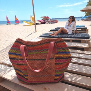 May include: A large, woven tote bag featuring horizontal stripes in vibrant colors including orange, blue, yellow, and black. The bag has two sturdy, smooth brown leather shoulder straps attached to the top, and a solid textured front pocket.