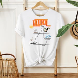 May include: White t-shirt with the word "LATINA" in orange block letters. The shirt features a map of Ecuador with illustrations of a sun, ship, toucan, and fish. The shirt is hanging on a wooden hanger.