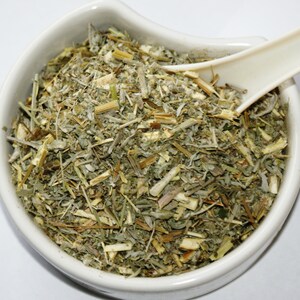 May include: A white ceramic bowl filled with dried herbs, featuring a mix of green, gray, and tan plant matter. A white spoon rests in the bowl, partially submerged in the herbs. The herbs appear to be a mix of leaves, stems, and other plant parts.