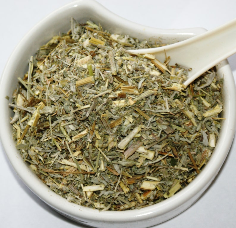 May include: A white ceramic bowl filled with dried herbs, featuring a mix of green, gray, and tan plant matter. A white spoon rests in the bowl, partially submerged in the herbs. The herbs appear to be a mix of leaves, stems, and other plant parts.