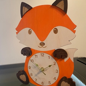 May include: A wooden fox clock with a brown tail and paws. The clock face is white with black numbers and gold hands. The fox is painted orange.