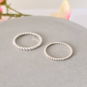 May include: Two silver beaded rings on a grey surface. The rings are made of small, round beads that are evenly spaced.