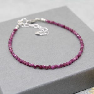 May include: A delicate bracelet featuring small, faceted, deep pink ruby beads strung on a thin wire. The piece includes a silver-toned metal lobster claw clasp and an adjustable chain extension with a small, flat, circular metal charm at the end.
