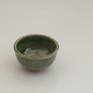 May include: A small, round ceramic bowl with a smooth, glossy finish in a muted sage green colour. The bowl features a slightly uneven, hand-crafted rim and a deep, concave interior that reflects light, showcasing the subtle variations in the glaze.
