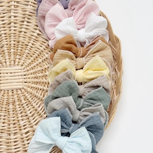 May include: A selection of pastel-coloured fabric bows, including pink, white, yellow, and blue, displayed in a woven basket. The bows have a delicate texture and design. These accessories are ideal for adding a touch of charm to any outfit or hairstyle.