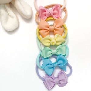 May include: A set of six pastel-coloured hair ties with matching bows. The bows are made of satin ribbon and are arranged in a rainbow order from pink to purple.