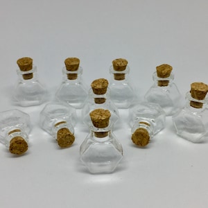 Pode incluir: Uma coleção de pequenas garrafas de vidro transparente com bases hexagonais e rolhas de cortiça. As garrafas estão dispostas sobre uma superfície branca, algumas em pé e outras deitadas. As rolhas são de cor castanho claro.