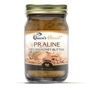 May include: A glass jar of Queen's Harvest Praline Pecan Honey Butter. The jar has a gold lid and a label with a bee logo. The label also says "Queen's Harvest" and "Praline Pecan Honey Butter".
