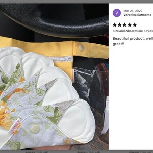 May include: Six reusable cloth menstrual pads with a floral print and white absorbent inserts. The pads are arranged in a stack and have a yellow fabric pouch behind them.