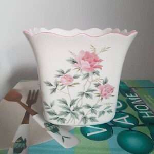 May include: White ceramic planter with a scalloped rim and pink floral design. The planter has a pink trim around the top edge.