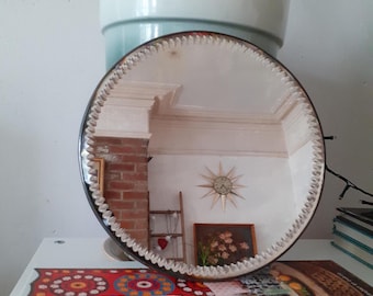May include: A round mirror with a decorative, textured edge. The mirror reflects a room with a brick wall, a starburst clock, and a painting. The mirror sits on a white surface with a book titled "The New Bohemians."