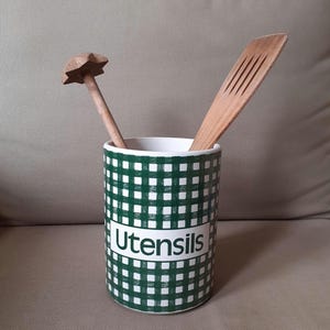 May include: A green and white checked utensil holder with the word "Utensils" printed on the front. It contains a wooden muddler with a star-shaped head and a slotted spatula. The holder is ceramic.
