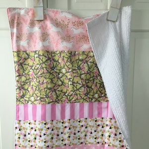 May include: A pink and green baby blanket with a white fleece backing. The blanket features a variety of patterns, including polka dots, stripes, and geometric designs.