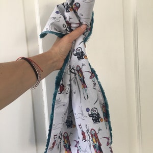 May include: A white fabric tea towel with a teal fuzzy border. The fabric features a repeating pattern of characters from the movie "The Nightmare Before Christmas".