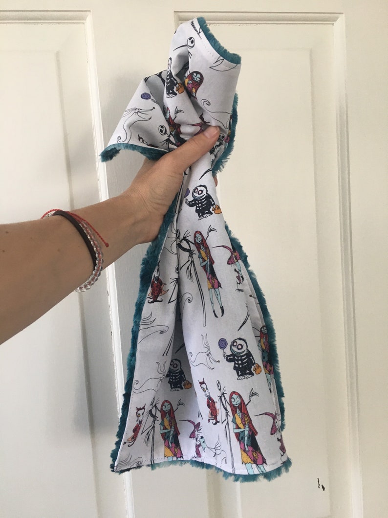 May include: A white fabric tea towel with a teal fuzzy border. The fabric features a repeating pattern of characters from the movie "The Nightmare Before Christmas".