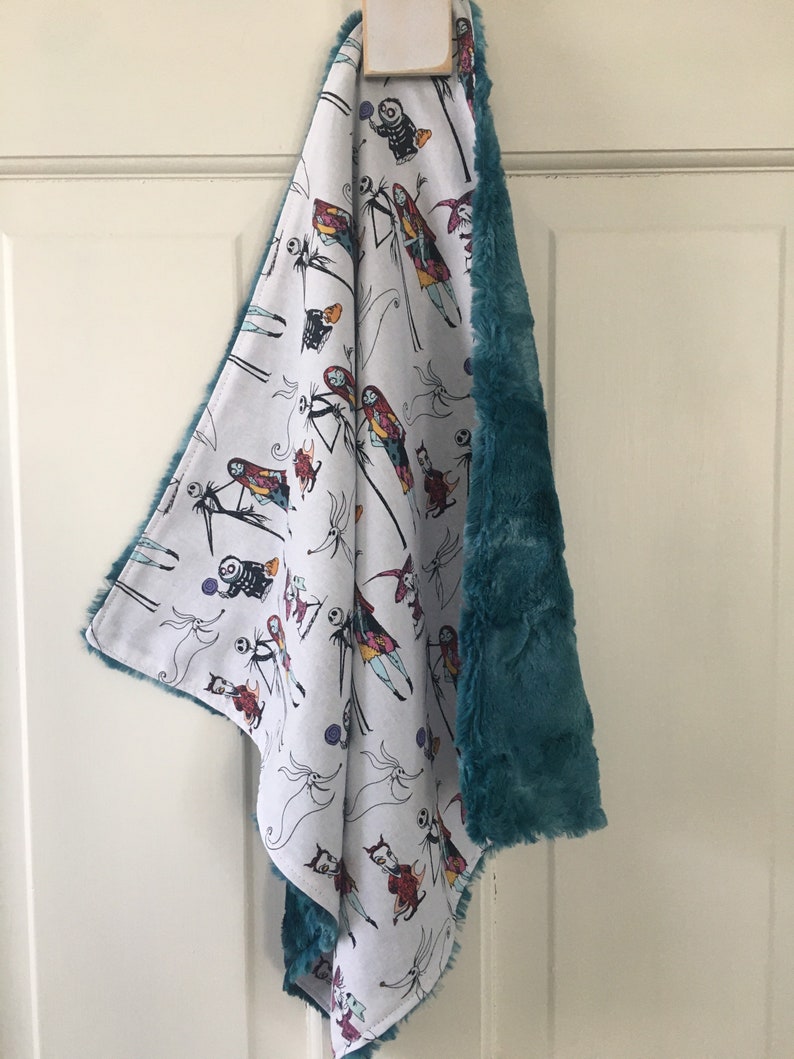 May include: A white and teal baby blanket featuring a Nightmare Before Christmas print. The blanket depicts various characters from the film, including Jack Skellington, Sally, and Oogie Boogie.