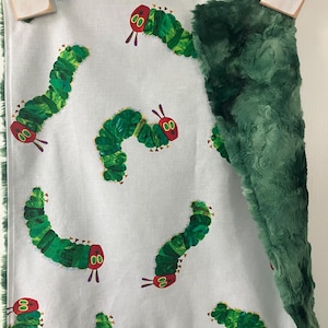May include: A green and white baby blanket with a green fuzzy side and a white side with a repeating pattern of green caterpillars with red heads. The blanket has a green fringe around the edges.