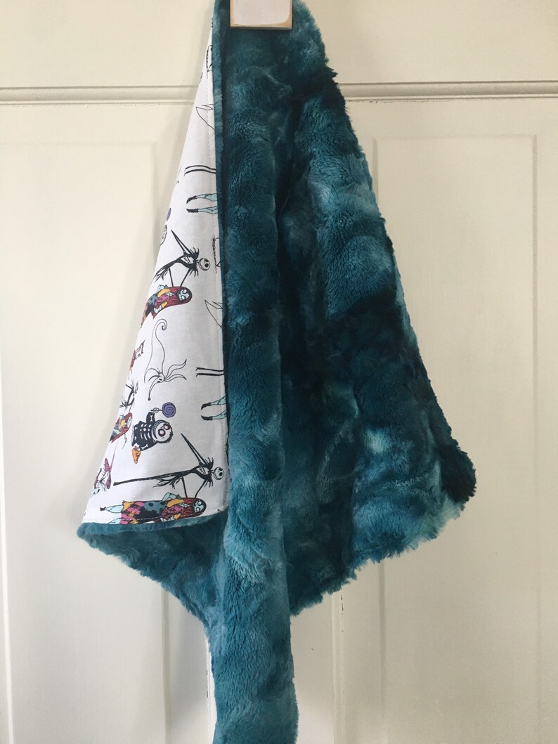 May include: A teal plush baby blanket with a white cotton side featuring a Nightmare Before Christmas print. The blanket is folded and hanging on a white door.