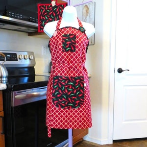 May include: A red apron with a white quatrefoil pattern, featuring a black pocket with a red and green chili pepper print. A matching square pot holder with the same chili pepper design hangs from the top of the display stand behind the apron.