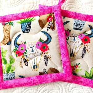 May include: Two pink and white quilted hot pads with a skull, cactus, and boot design. The hot pads have a pink border and a white background with a colorful floral design.