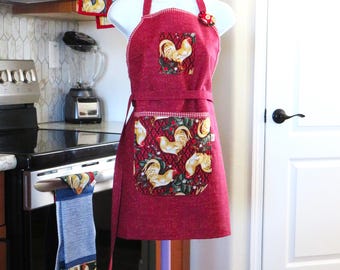 May include: A red apron featuring a rooster print on the chest and a larger pocket with a repeating rooster pattern. The apron has a white neck strap and waist ties. A small tag near the neck reads Size Fits All. The apron is displayed on a stand.