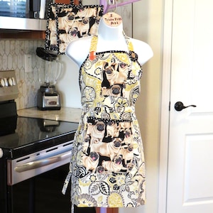 May include: An apron and matching pot holder set featuring a repeating pattern of pug dogs on a cream background with yellow and black floral accents. The apron is displayed on a mannequin in a kitchen setting.