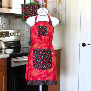 May include: A red apron with a black and red chili pepper print on the pockets and hem, featuring a paisley pattern on the main body. A matching square potholder hangs nearby. A white tag at the top reads Aprons ONE SIZE FITS ALL.