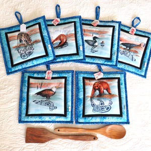 May include: A set of six square, quilted pot holders with blue tie-dye borders and black trim. Each features a different animal illustration: wolf, duck, loon, whale, eagle, and bear. Two wooden cooking utensils are below.
