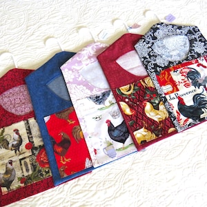 May include: Five fabric garment covers with various rooster and floral patterns. The covers are red, blue, and white, with a white hanger at the top. The covers are made of fabric and are designed to protect clothing.
