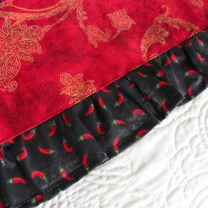 May include: A textile item featuring a vibrant red fabric with a gold-toned floral paisley pattern, finished with a black ruffled border decorated with a repeating print of small red chili peppers, showcasing a distinct contrast in color and texture.
