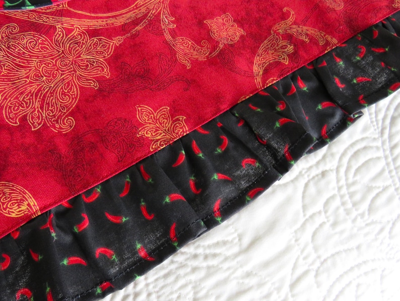 May include: A textile item featuring a vibrant red fabric with a gold-toned floral paisley pattern, finished with a black ruffled border decorated with a repeating print of small red chili peppers, showcasing a distinct contrast in color and texture.