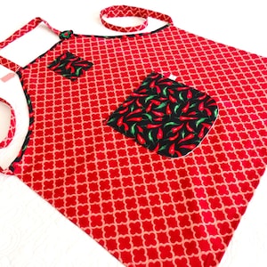 May include: A red apron featuring a repeating light-colored clover pattern, accented with black trim and two pockets. The pockets are decorated with a vibrant print of red and green chili peppers on a black background, complemented by matching straps.