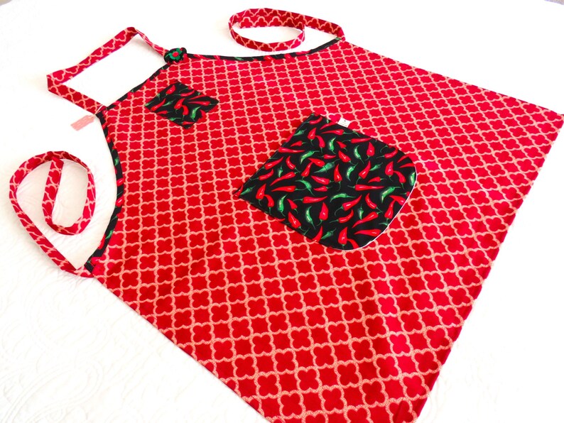 May include: A red apron featuring a repeating light-colored clover pattern, accented with black trim and two pockets. The pockets are decorated with a vibrant print of red and green chili peppers on a black background, complemented by matching straps.