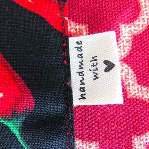 May include: A fabric label with the text "handmade with" and a black heart icon, sewn onto a seam between a black fabric featuring red chili peppers and a red fabric with a white geometric lattice pattern. The stitching is visible in red thread.