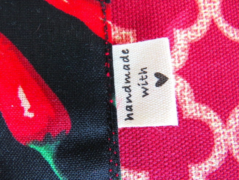 May include: A fabric label with the text "handmade with" and a black heart icon, sewn onto a seam between a black fabric featuring red chili peppers and a red fabric with a white geometric lattice pattern. The stitching is visible in red thread.