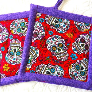 May include: Two purple and red quilted pot holders with a sugar skull pattern. The pot holders have a purple border and a red background with white sugar skulls and colourful flowers.