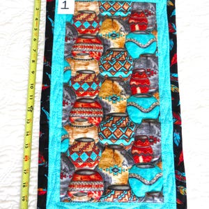 May include: A rectangular wall hanging featuring a repeating pattern of colorful pottery. The central panel is bordered by a turquoise frame and a black fabric with red, yellow, and blue feathers. The wall hanging is approximately 21 inches long.