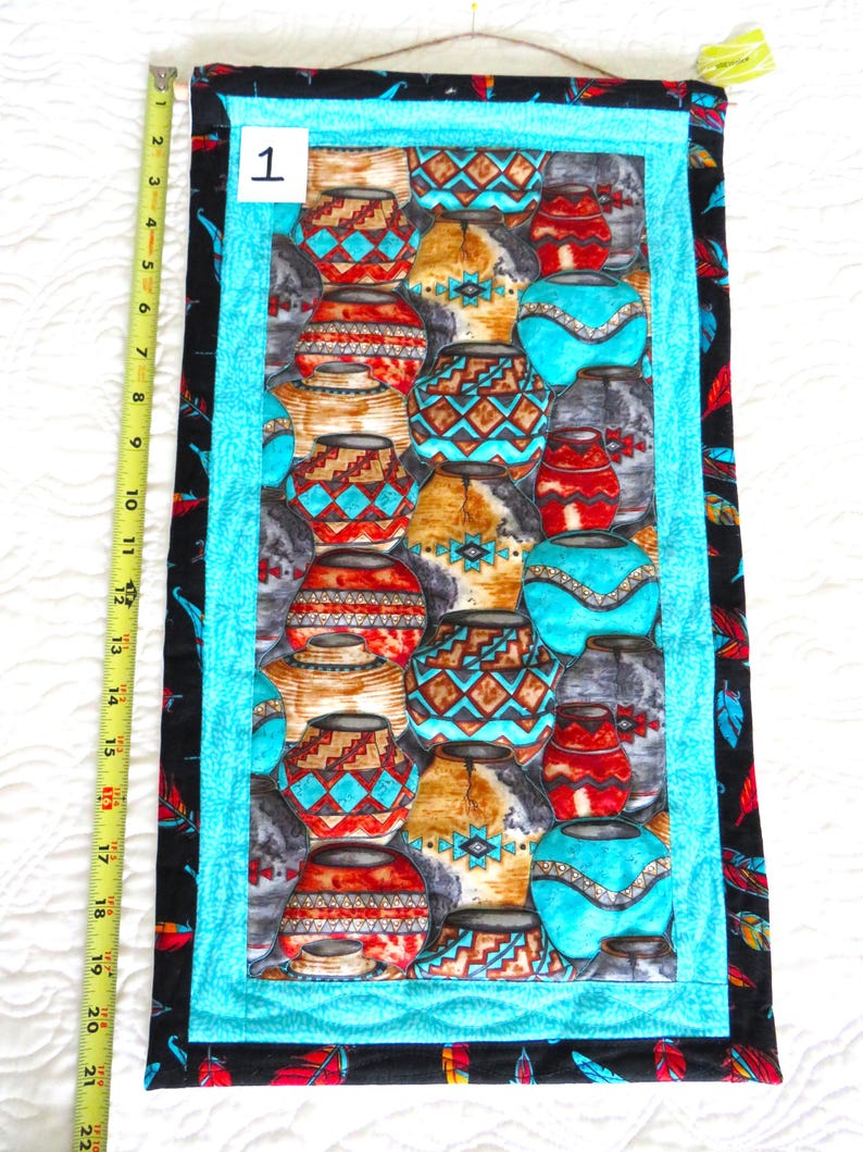 May include: A rectangular wall hanging featuring a repeating pattern of colorful pottery. The central panel is bordered by a turquoise frame and a black fabric with red, yellow, and blue feathers. The wall hanging is approximately 21 inches long.