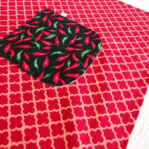 May include: A red fabric piece featuring a repeating light-colored quatrefoil pattern. Attached to the surface is a black square pocket with rounded corners, decorated with a vibrant print of numerous small red and green chili peppers.