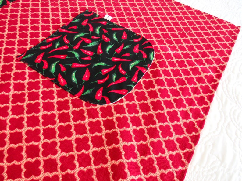 May include: A red fabric piece featuring a repeating light-colored quatrefoil pattern. Attached to the surface is a black square pocket with rounded corners, decorated with a vibrant print of numerous small red and green chili peppers.